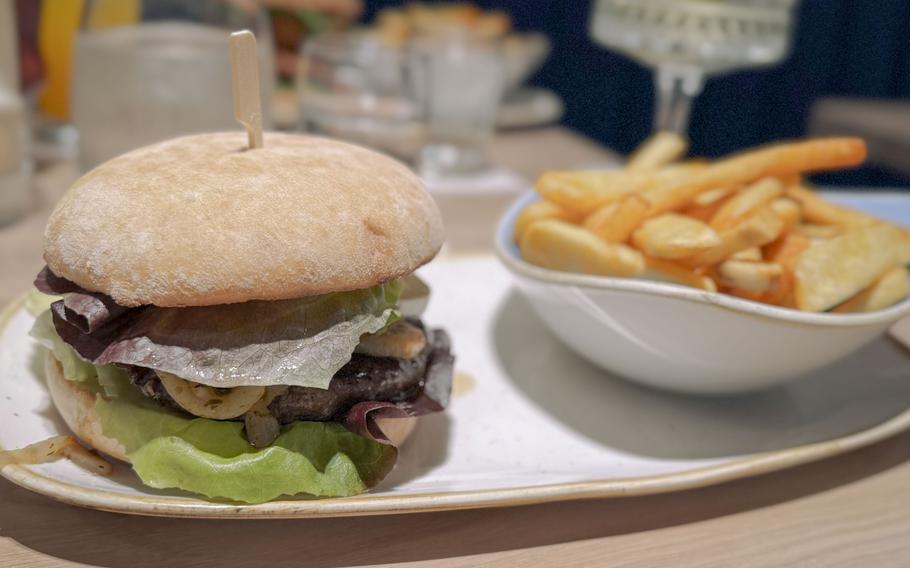 Gourmet beef burger with lettuce and cured meat on a soft bun, served with crispy fries in a white bowl om the side.