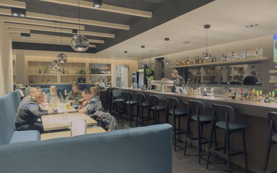 Interior dining area with customers seated at tables and a modern bar with pendant lights, shelving with bottles, and contemporary teal upholstered seating.