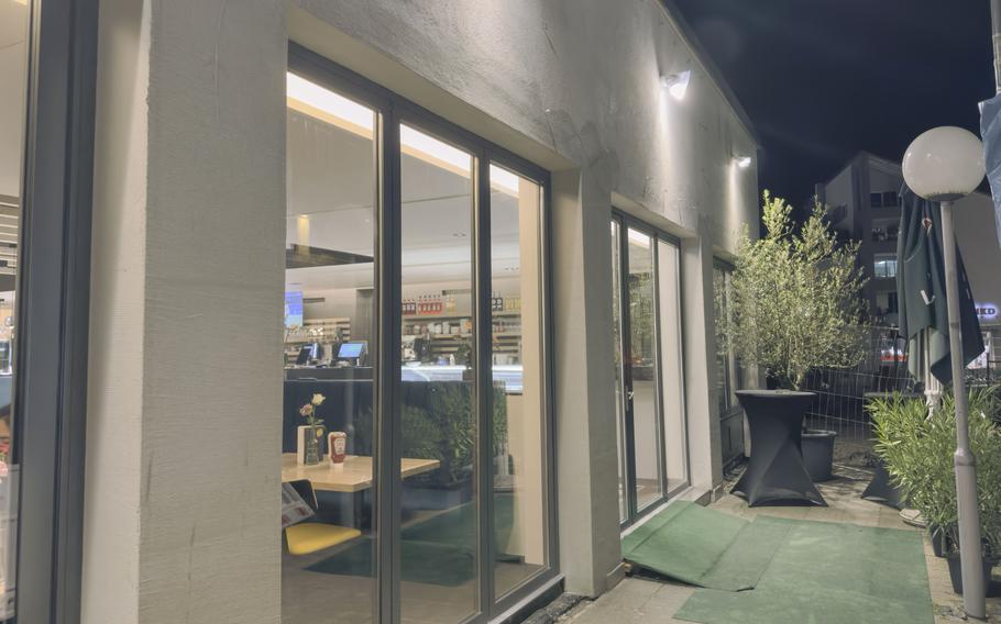 Exterior of Schwan Mediterranean Diner at night, showing modern storefront with large glass windows, illuminated facade, green artificial turf entrance and greenery.