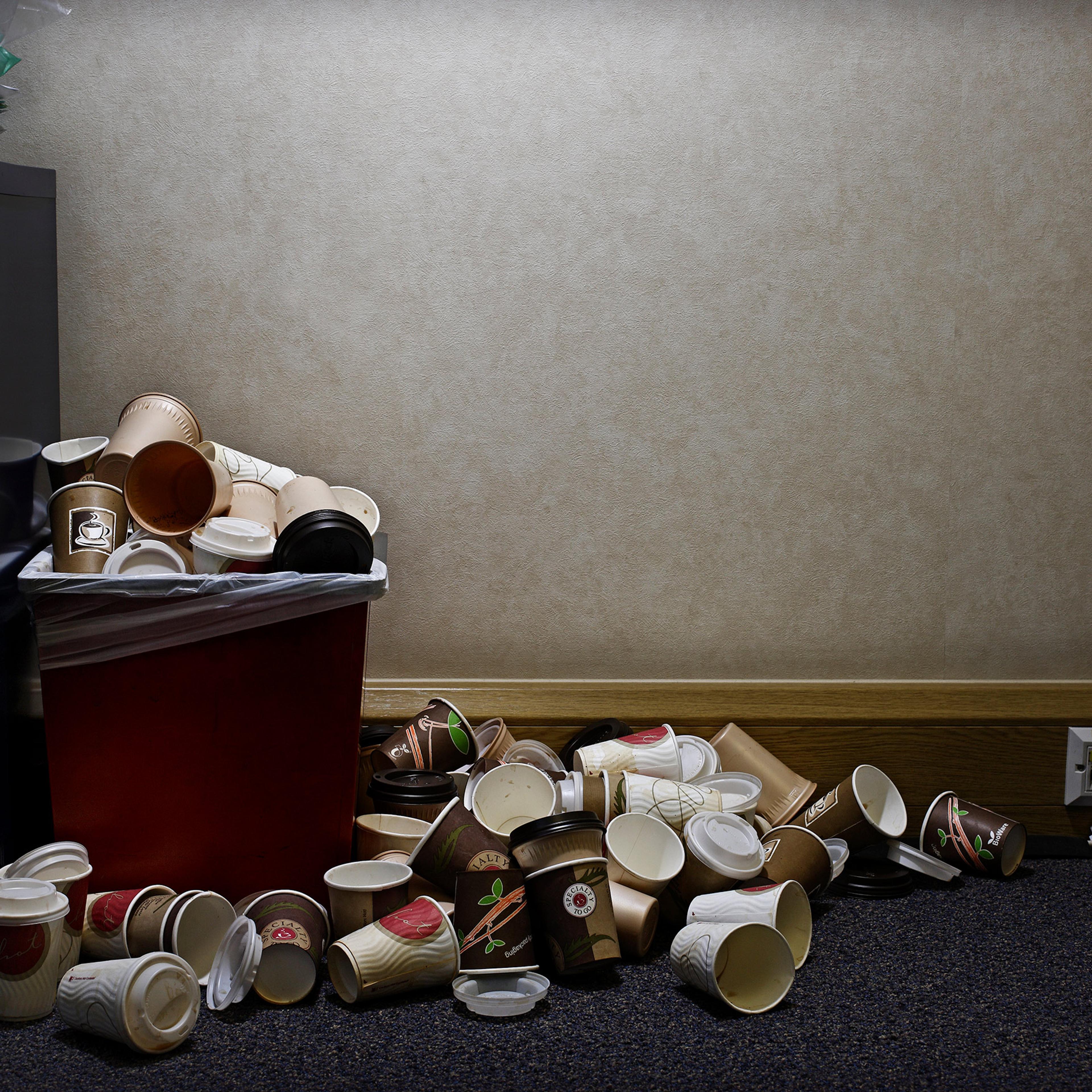 A red bin overflowing with paper coffee cups next to a wall with powerpoints in an office setting.