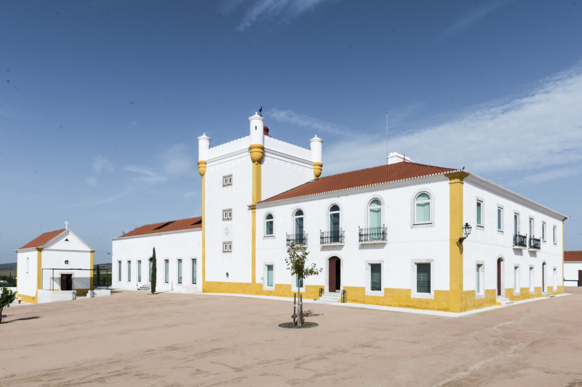 White historic building with a central tower, red roof, and blue sky backdrop in a spacious courtyard.