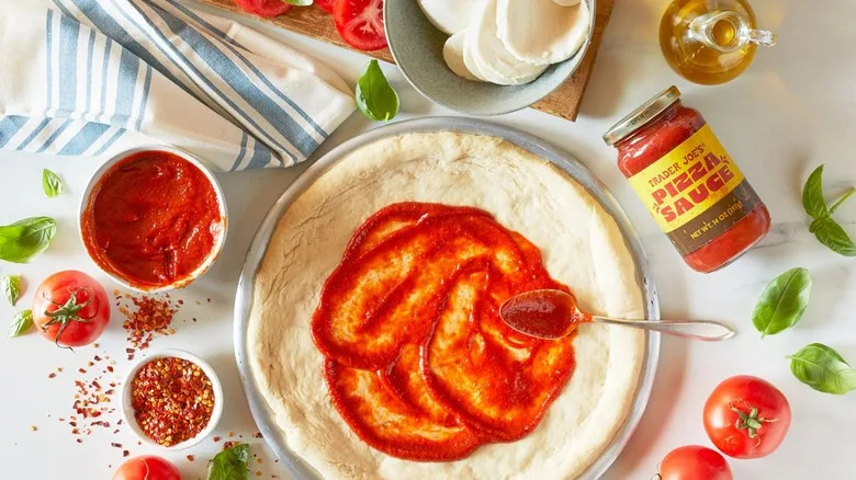 Trader Joe's pizza sauce being spread on an unbaked pizza crust surrounded by other pizza ingredients