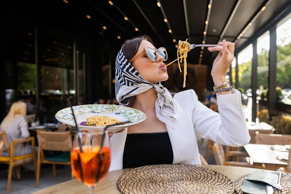 A fashionable woman eating spaghetti and drinking aperol at an elegant outdoor restaurant setting. Stylish attire and lively dining atmosphere.