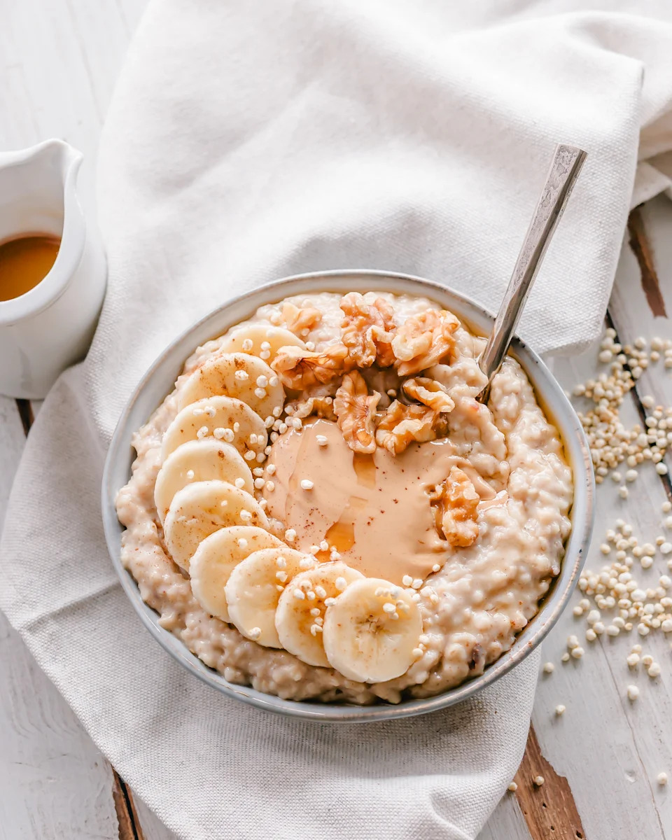 Bowl of creamy cooked oatmeal topped with banana, cashew butter, walnuts, quinoa puffs, syrup and cinnamon