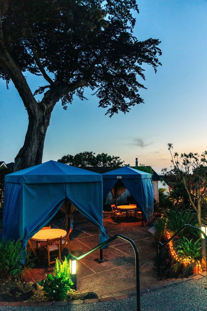 Blue cabanas with round tables and chairs are lit warmly at dusk, surrounded by plants and string lights under a large tree.