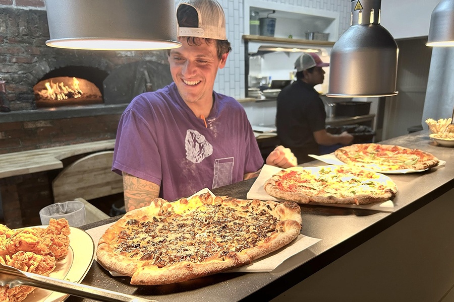A chef in a t-shirt and backwards baseball cap smiles behind a lineup of pizzas, with a pizza oven firing behind him.