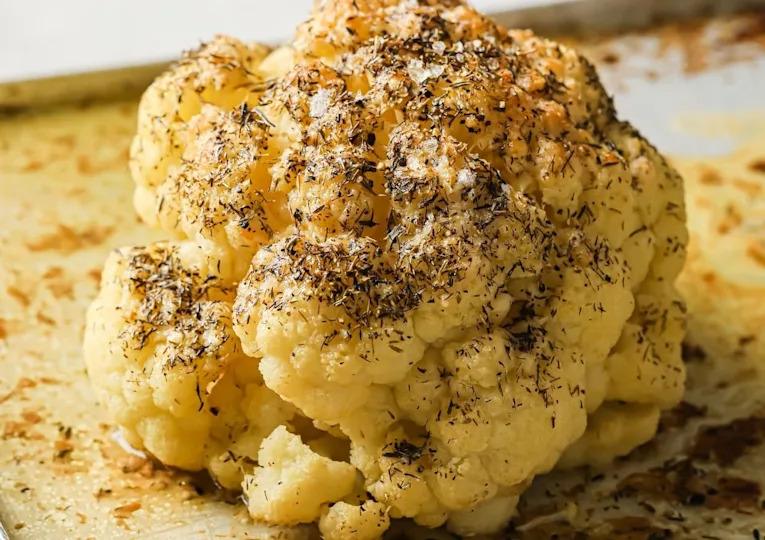 A roasted whole cauliflower head topped with herbs and seasoning on a baking tray.