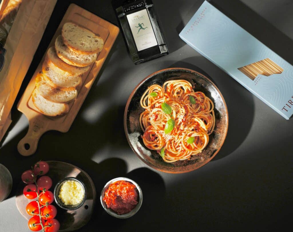Celebrate National Pasta Day with a bowl of spaghetti topped with tomato sauce and basil, served alongside sliced bread, cherry tomatoes, grated cheese, and a jar of sauce—all set on a dark table with packaged pasta and olive oil.