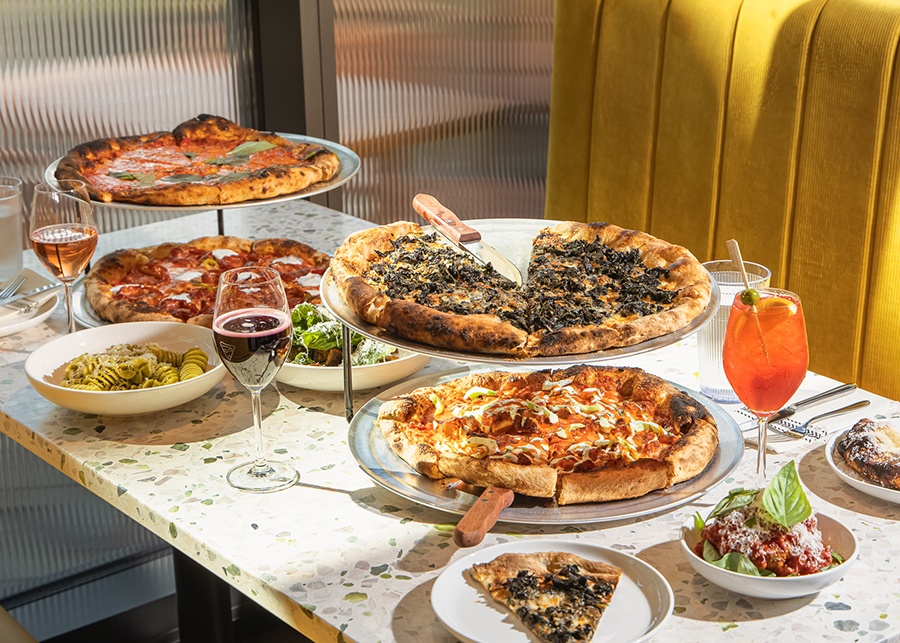 Four pizzas with puffy, lightly charred crusts sit on a terrazzo table at a restaurant with cocktails, wine, appetizers, and a bit of a mustard-yellow banquette peeking through.