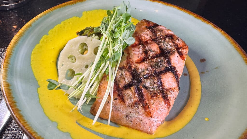 a grilled fish dish with yellow sauce, lotus root, and green sprout garnish