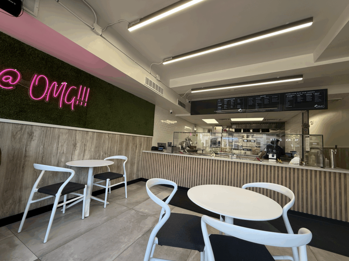 A restaurant with a sign on the wall that reads “OMG” and several small tables, as well as an order counter with build-your-own style options and a menu overhead.