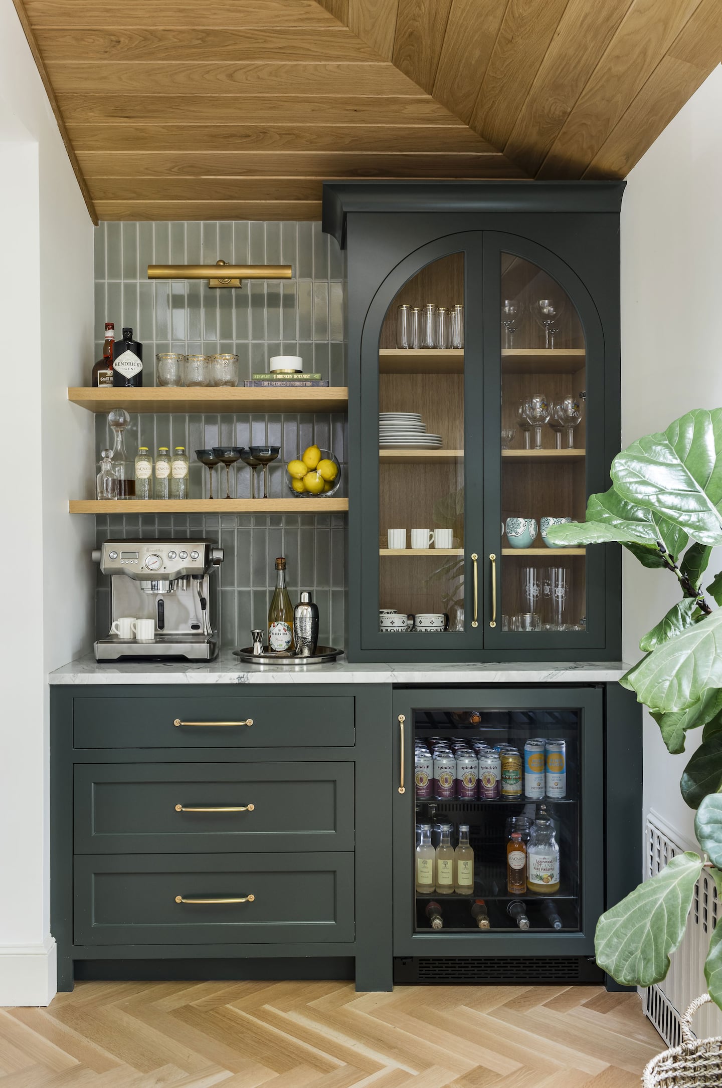 A bar cabinet with dark green cabinets. On the right, there's a glass front cabinet. Below that there's a glass front refrigerator. ON the left, the lower cabinets have draws and the counter above holds an espresso machine. Above that, there are open shelves with wine glasses.
