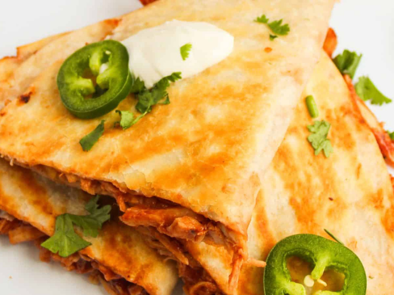 Close-up of chicken quesadillas topped with sour cream, sliced jalapeños, and fresh cilantro on a white plate.