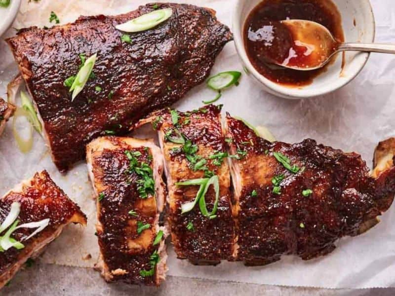 Sliced barbecued ribs topped with green onions and herbs, served on parchment paper with a bowl of sauce and a spoon.