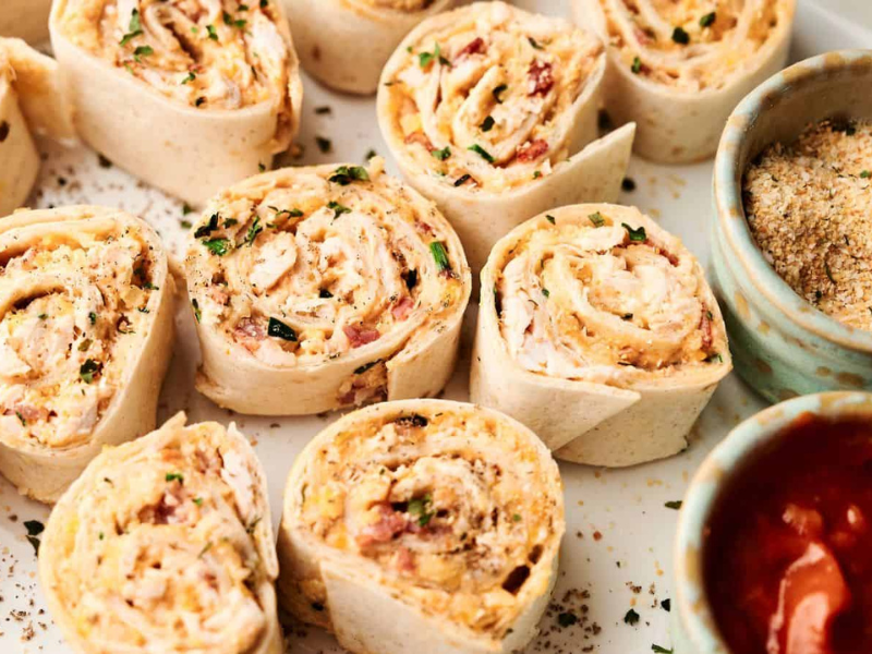 Close-up of several tortilla pinwheels filled with a creamy mixture, garnished with herbs, next to small bowls of breadcrumbs and red dipping sauce.
