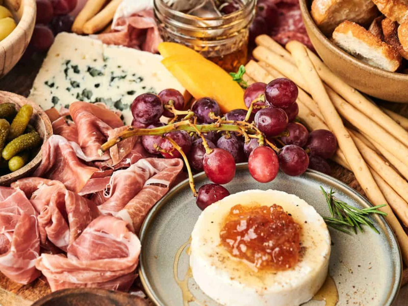 A charcuterie board with grapes, prosciutto, breadsticks, cornichons, blue cheese, a cheese round topped with marmalade, and a bowl of sliced bread.