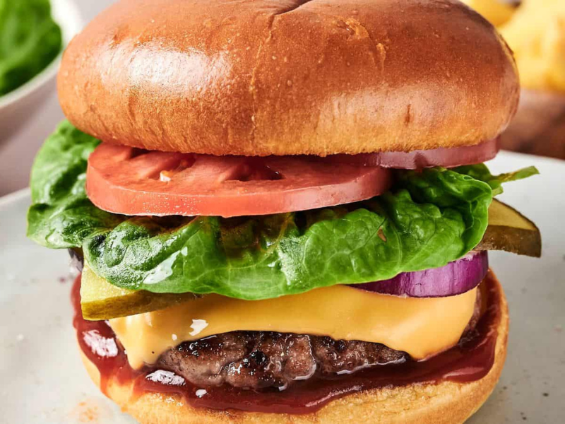 A cheeseburger with lettuce, tomato, pickles, red onion, cheddar cheese, and ketchup in a brioche bun on a white plate.