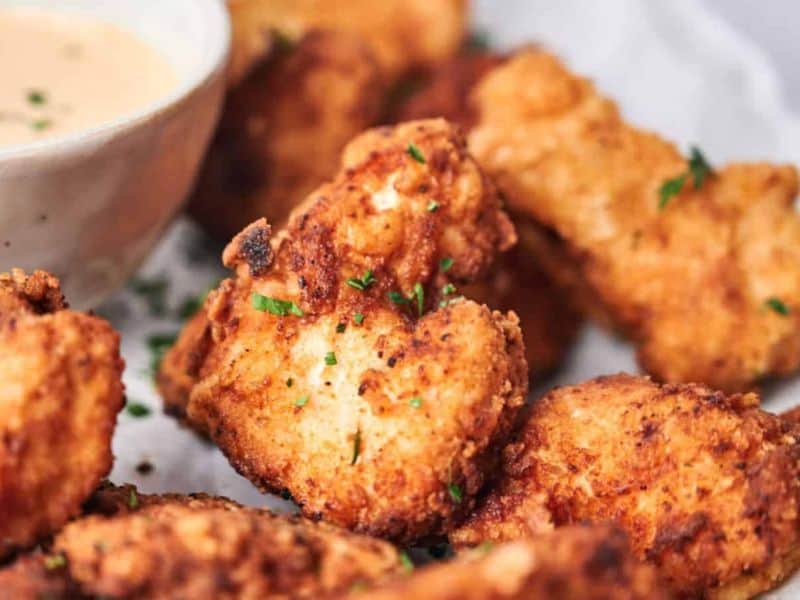 Close-up of golden-brown fried chicken pieces garnished with herbs, with a bowl of dipping sauce in the background.