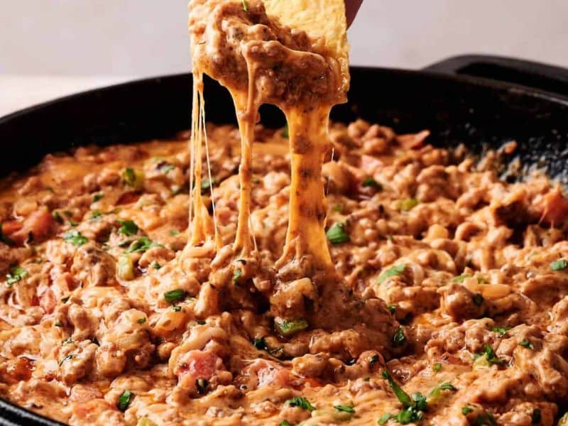 A tortilla chip is dipped into a skillet of cheesy ground beef dip, with melted cheese stretching between the chip and the dip.