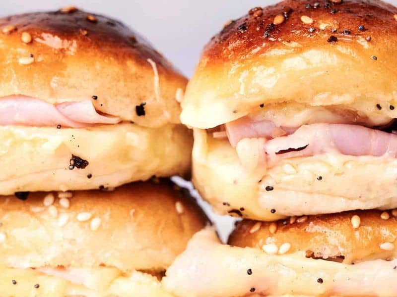 Close-up of stacked ham and cheese sliders on sesame seed buns, showing melted cheese and slices of ham inside golden, glossy bread.
