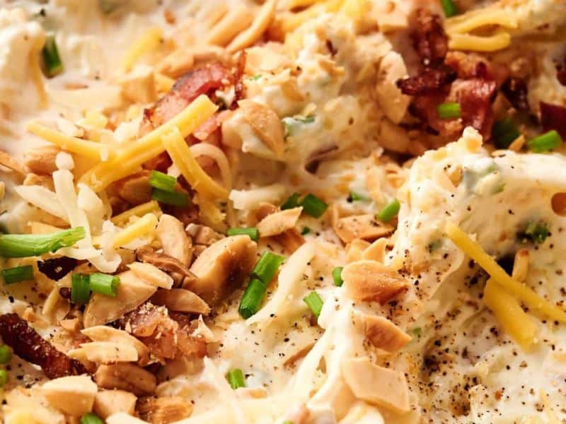 Close-up of a creamy dip topped with shredded cheese, chopped chives, sliced almonds, bacon pieces, and ground black pepper.