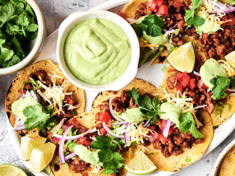 Plate of ground beef tacos topped with shredded cheese, onions, tomatoes, cilantro, and avocado sauce, served with lime wedges and a bowl of green sauce on the side.