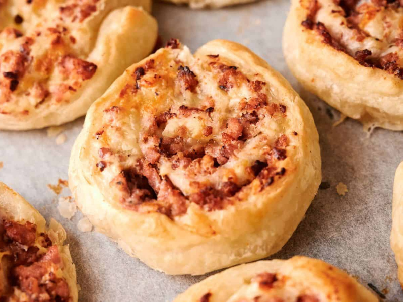 Close-up of baked pinwheel pastries with a golden, flaky crust and a visible savory filling, arranged on parchment paper.