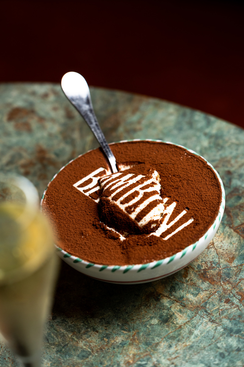 a huge tiramisu in a bowl, with 'bambini' written across the top in cocoa powder and a spoon plunged into the middle