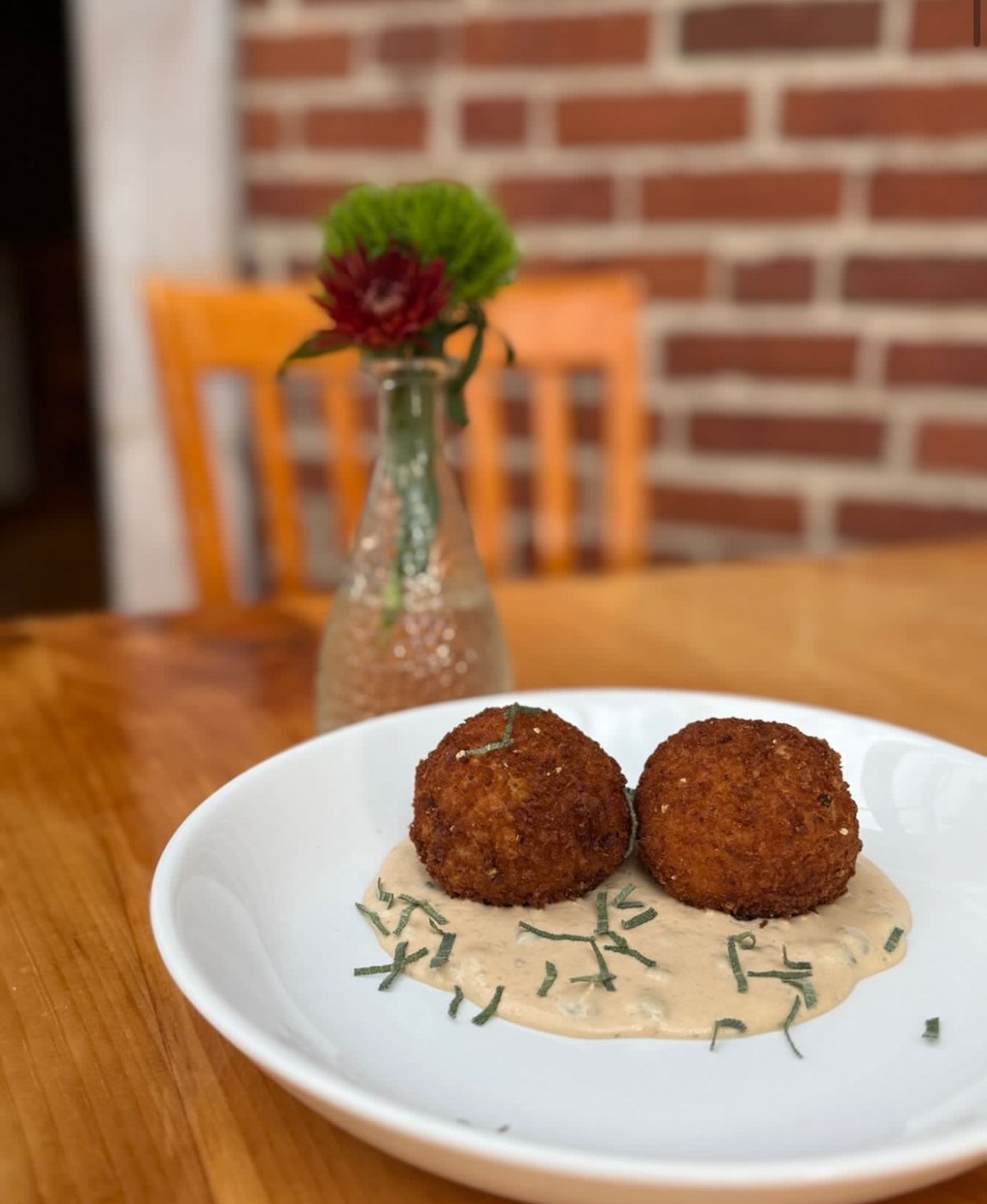 Two breaded and fried balls served on a creamy sauce with herb garnish, on a white plate placed on a wooden table; a vase with flowers and a brick wall are in the background.