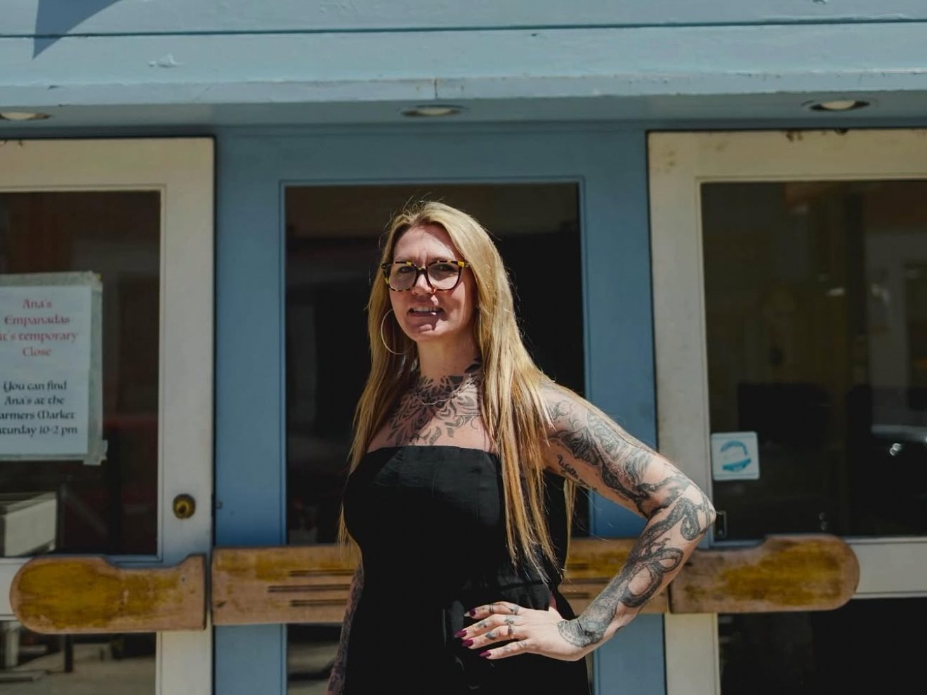 A woman with long blonde hair, glasses, and visible tattoos stands in front of a glass double door, wearing a black sleeveless dress.