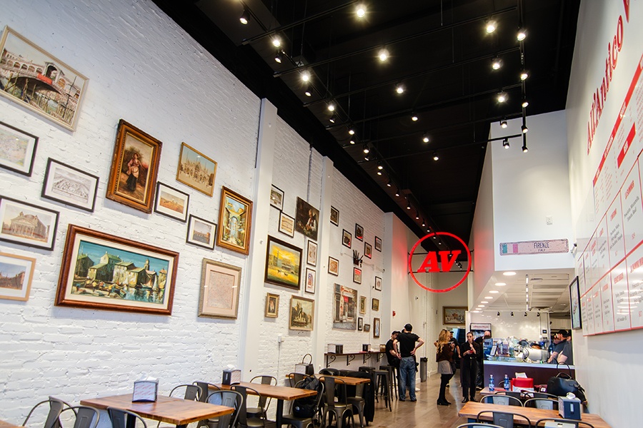 Interior of a small casual counter-service restaurant with pictures of Italy, a sign with an Italian flag that says Firenze (Florence), and neon red signage with the letters A and V in a circle, short for the restaurant's name, All'Antico Vinaio.