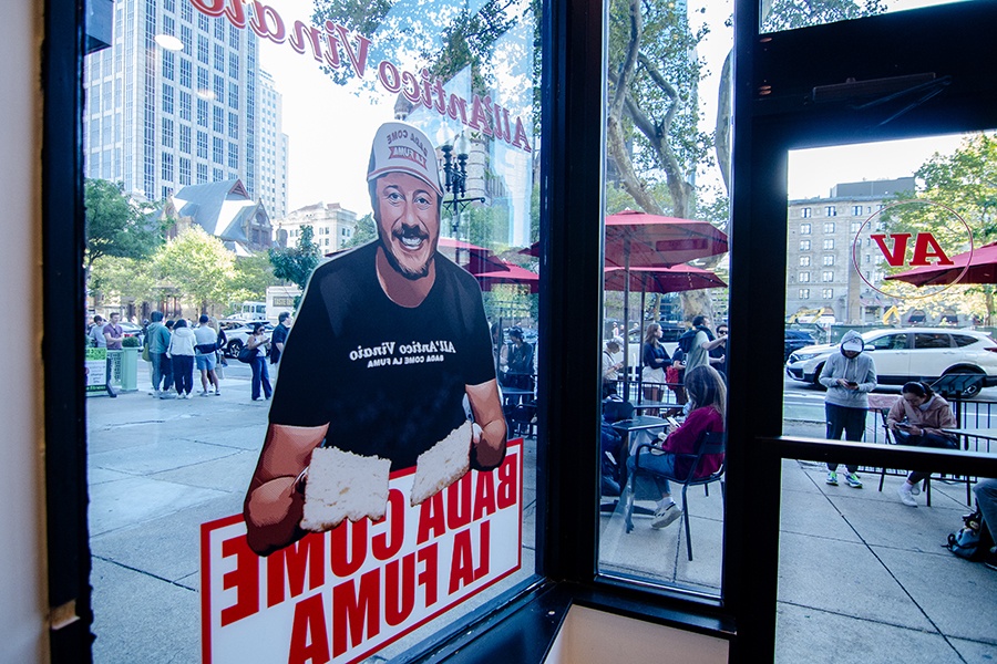 Photographed from inside a restaurant, a line begins to form outside. A window of the restaurant features a large decal of the owner holding fresh bread.