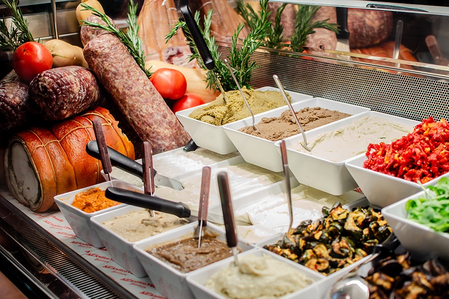 A refrigerated case at an Italian counter-service restaurant holds various meats, tomatoes, rosemary, and condiments.