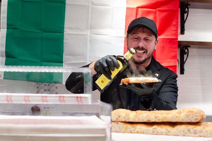 A man behind the counter at a casual restaurant pours olive oil onto hot flatbread. An Italian flag hangs behind him.