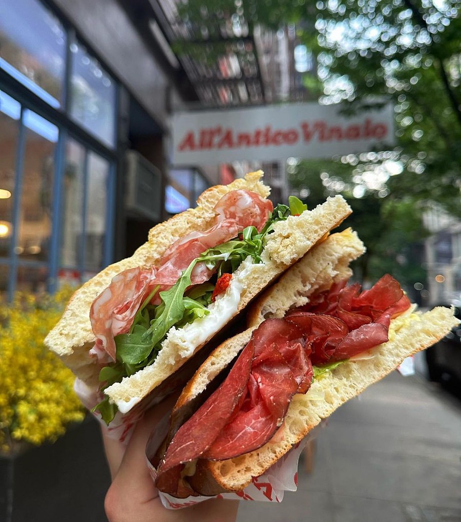 A hand holds up a big sandwich on Italian flatbread in front of a sign that says All'Antico Vinaio.