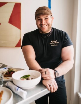 a white man wearing a brown hat and black tshirt with the words piccola cucina stands smiling near a white bowl of prepared food
