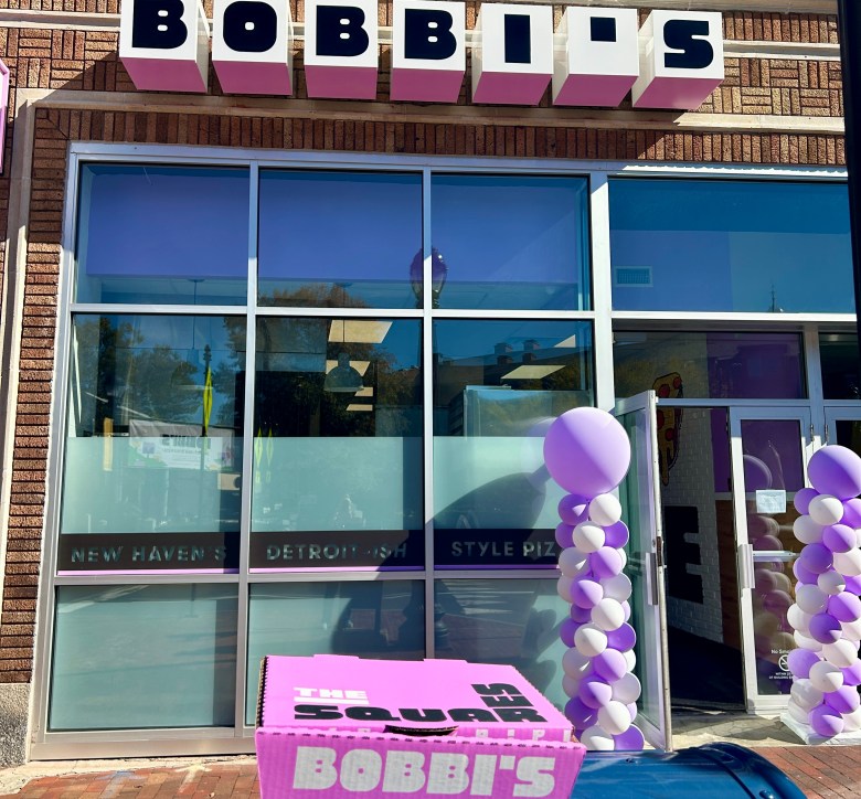 Store exterior that reads Bobbi's with purple and white balloons and purple pizza box in foreground.