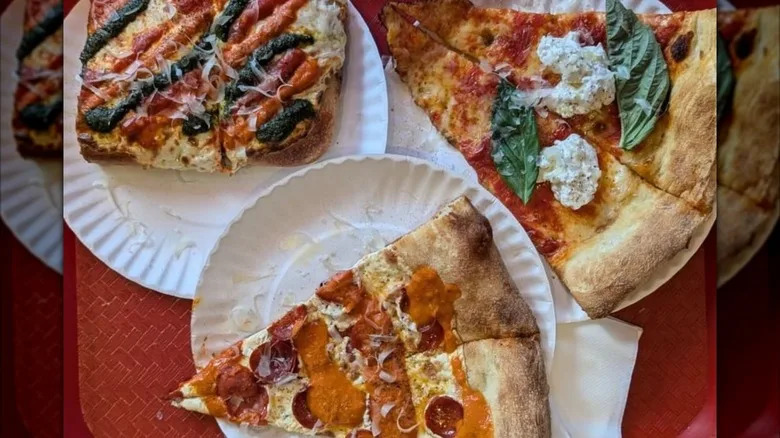A tray carrying three different flavors on pizza slices on white paper plates from Andrew Bellucci's Pizzeria