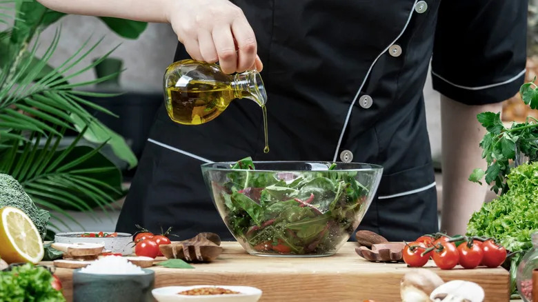 Someone in a chef coat drizzling olive oil over a bowl of mixed greens resting on a counter that is covered in fresh vegetables, herbs, and greens.