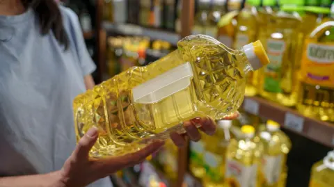 Getty Images Close-up of a shopper holding a bottle of cooking oil in a supermarket aisle, focusing on food choices, daily shopping, and consumer lifestyle.