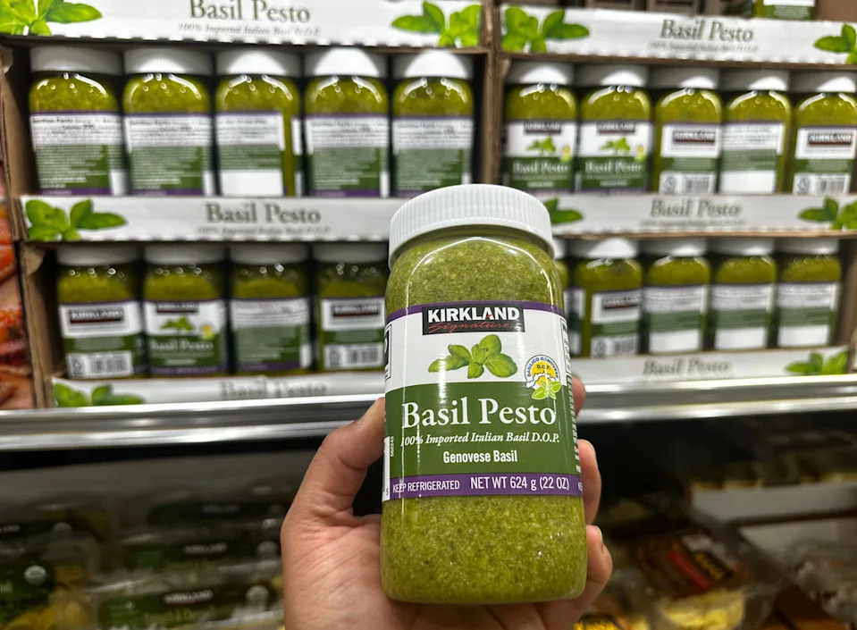 Hand holding Kirkland Signature basil pesto in front of aisle of pesto in Costco