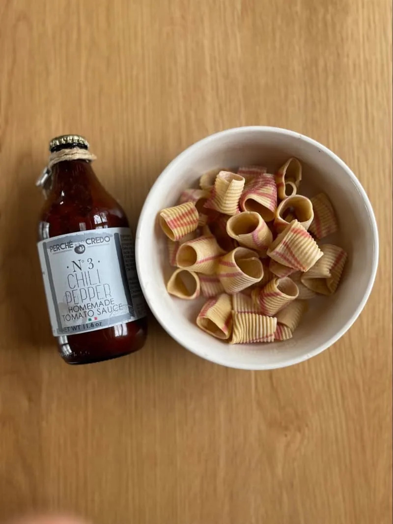 Striped pasta in a white bowl next to a bottle of chili pepper tomato sauce on a wooden surface.