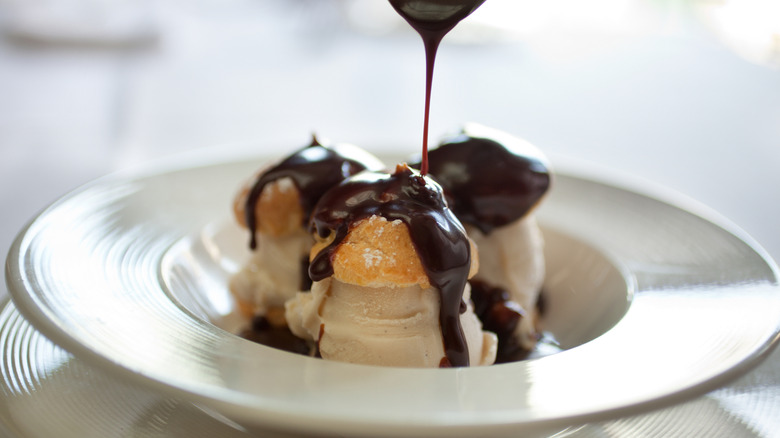 Three profiteroles in a white bowl with chocolate sauce being drizzled on top