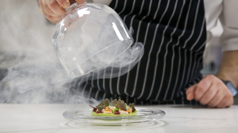 Chef lifting glass lid of dish, producing vapor