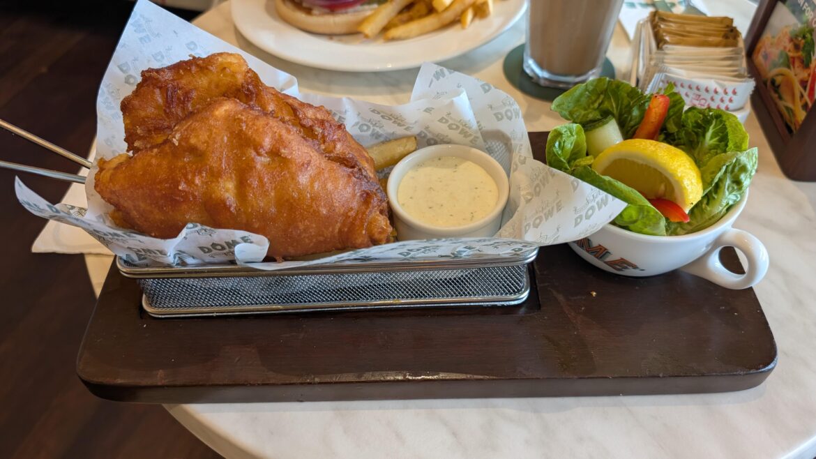 Fish and chips in a fryer basket, salad in a teacup, all on a wooden board