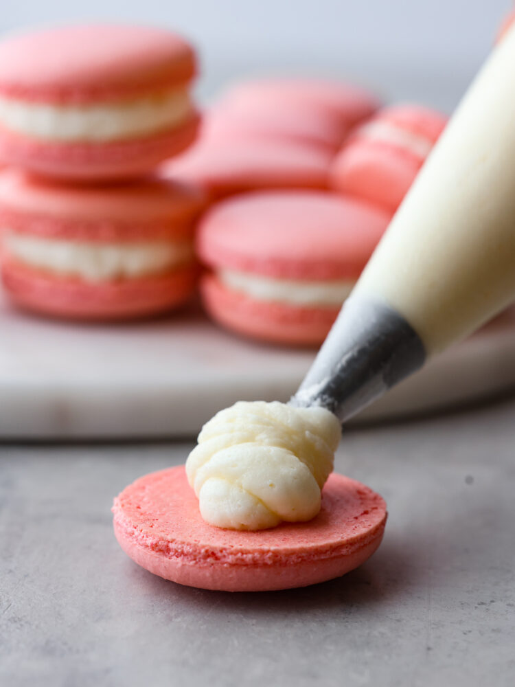 This French macaron cookie recipe makes the perfect macaron every time! I share all of my tips and tricks to get the perfect feet on your macaron, and a delicate and airy cookie with a shiny smooth shell. You are going to love making these fun and elegant cookies at home! A photo showing the filling being piped onto one half of the cookie.