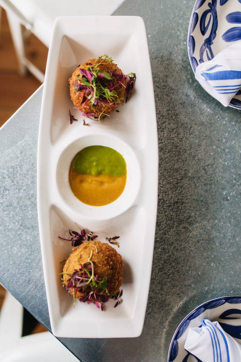 a long white plate with two orange rice balls and an orange and green sauce between them sits on a table at piccola cucina