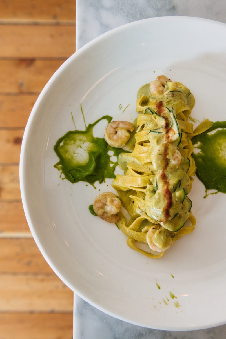 a round white plate containing a scoop of pasta flanked by shrimp and sliced zucchini sits on a table at piccola cucina