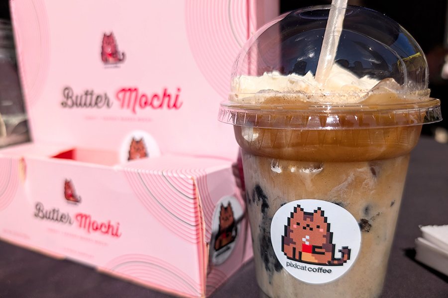 A latte with whipped cream is served in a plastic cup with a sticker featuring a pixelated cat, next to a pink box that says butter mochi.