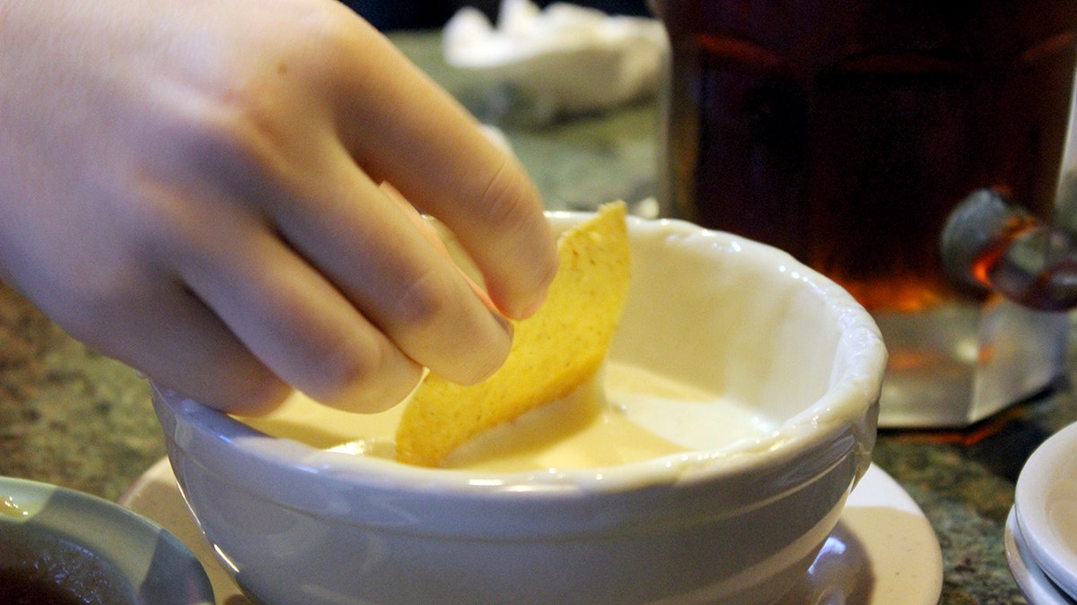 A hand holds a tortilla chip that is being dipped with queso from a white bowl.